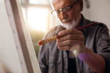 An elderly man paints on a canvas, paintbrush in hand, demonstrating how he's living with Alzheimer's.